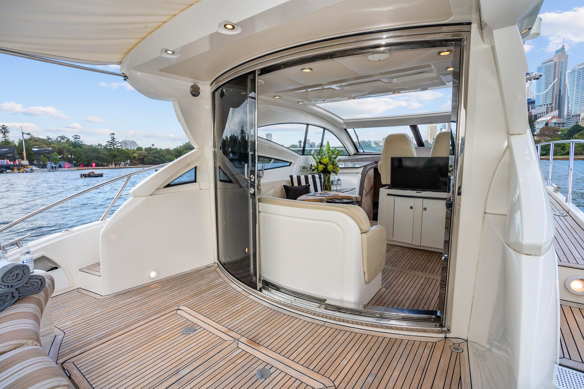 Experience the Birchgrove’s luxury yacht deck with wood flooring and a lounge area. Sliding glass doors reveal a modern interior with seating, a table, and flowers, all set against scenic water views and city buildings.