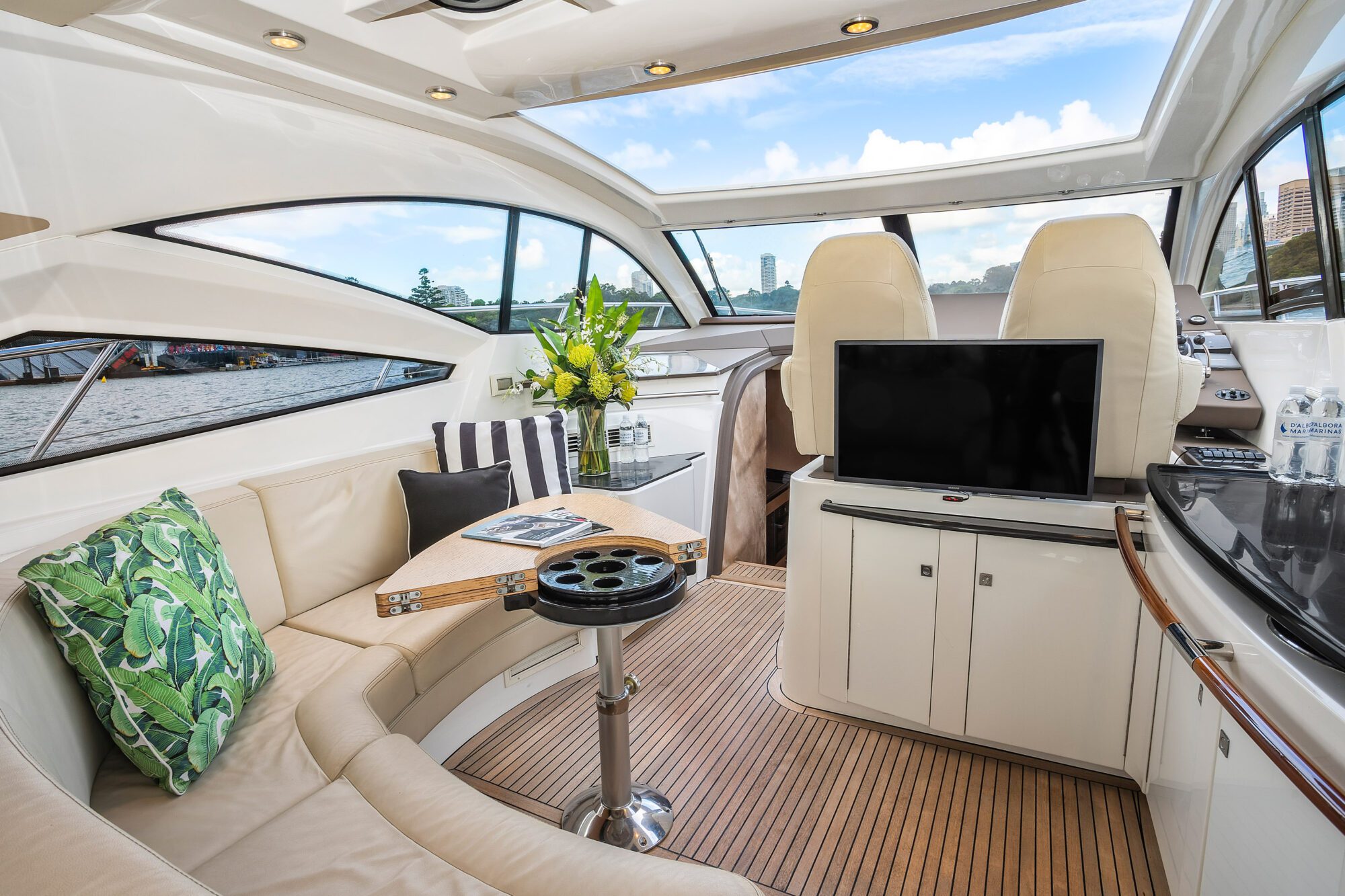 The Birchgrove luxury yacht interior features cream leather seating, decorative pillows, a wooden table with a flower vase, a TV, and large windows offering scenic views of the water and sky.