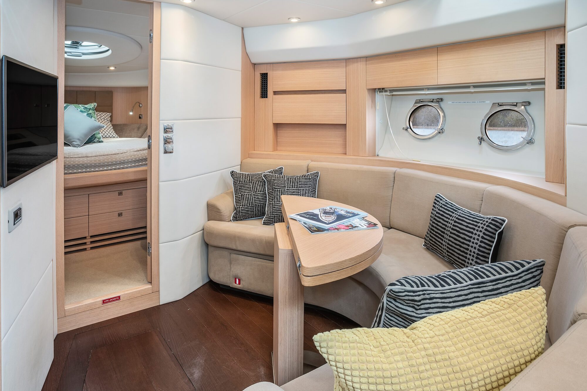 The Birchgrove features an elegant yacht interior with a curved beige sofa, patterned cushions, wooden table, round windows, and a cozy bedroom with pillows and sunlight streaming through a ceiling hatch.