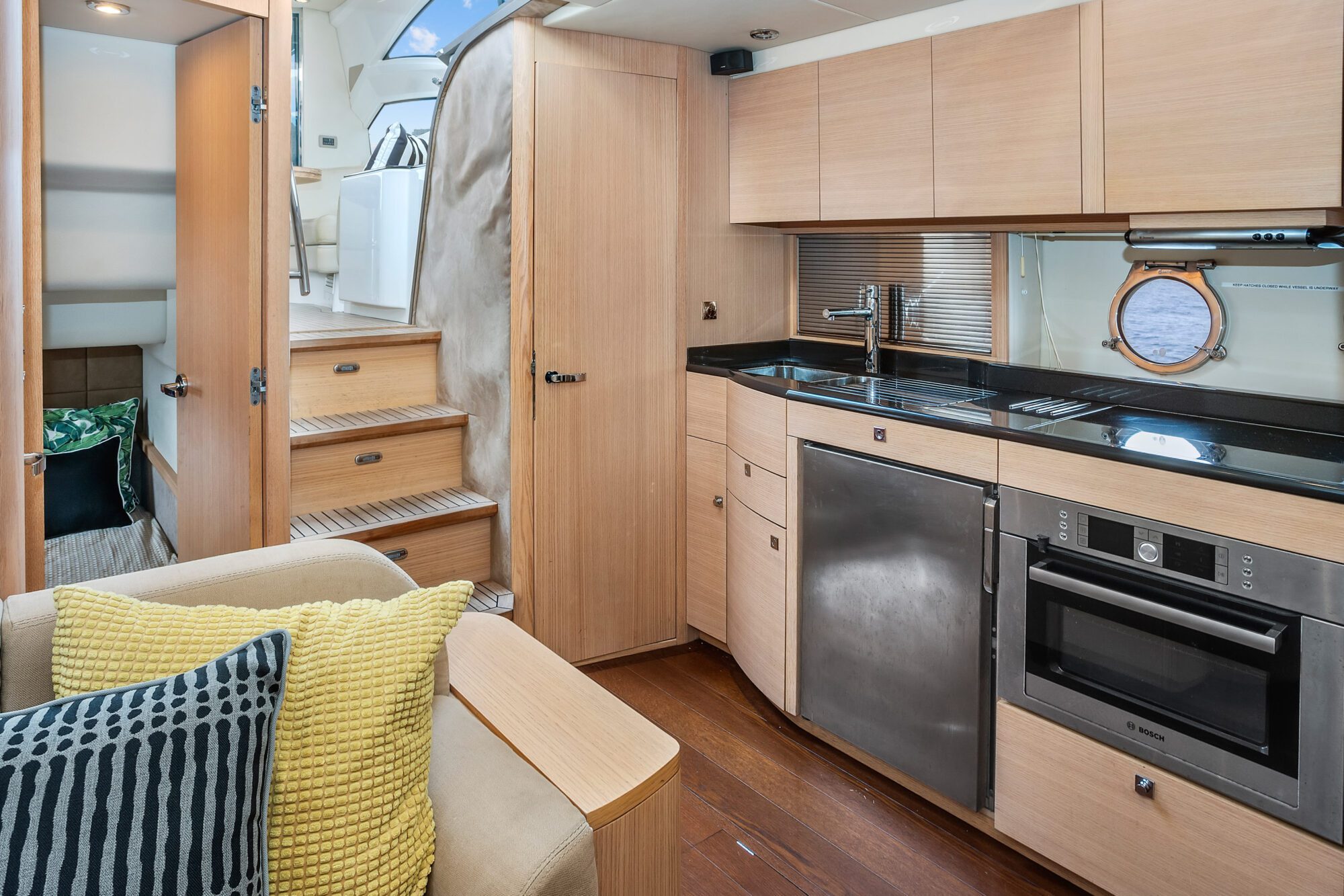 The Birchgrove features a modern yacht kitchen with light wood cabinets, stainless steel sink, oven, and dishwasher. Stairs lead to a door, with a sofa and yellow and black pillows nearby. A round window offers a view of the water.