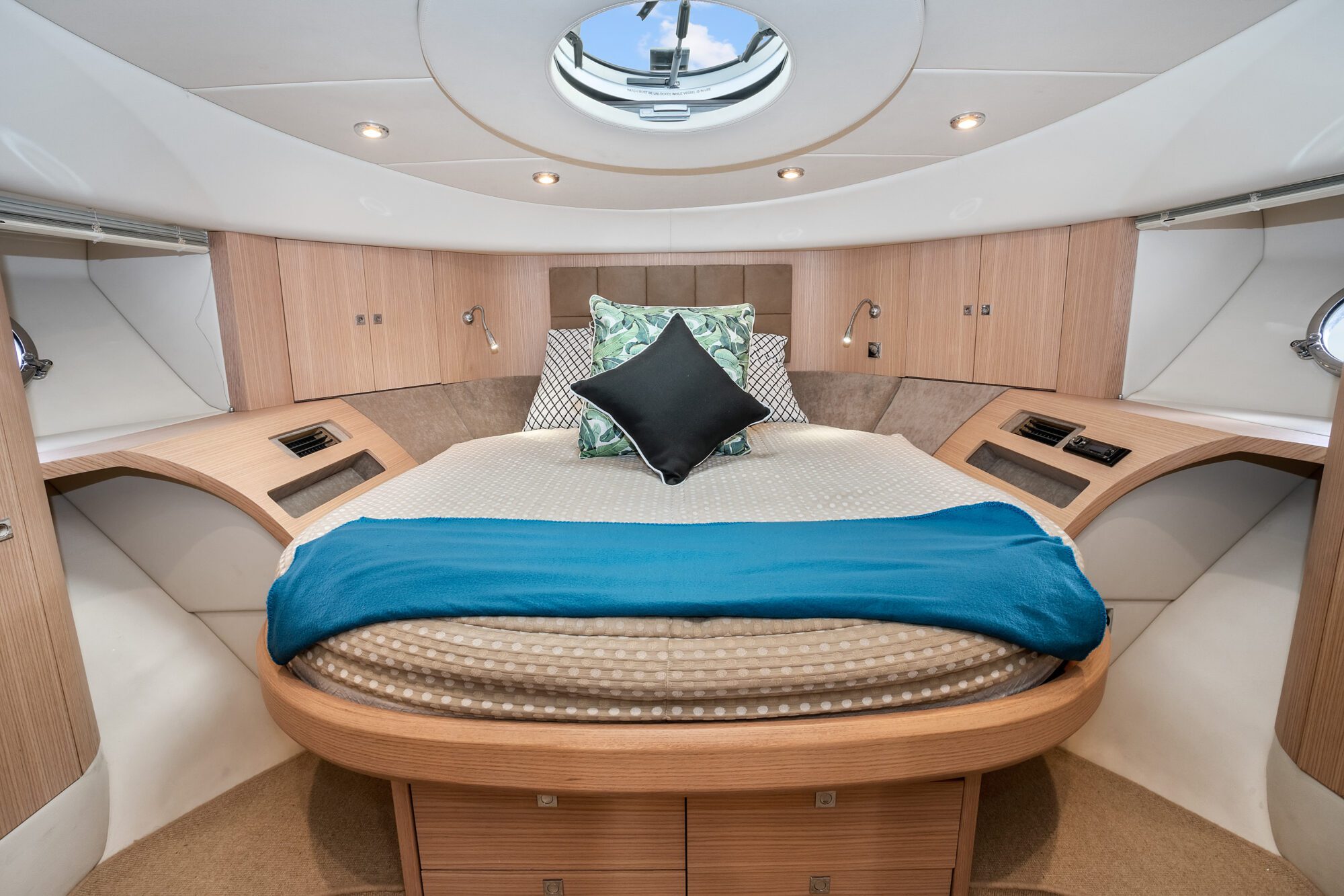 The Birchgrove features a modern boat cabin bedroom with a rounded bed, wood paneling, blue blanket, decorative pillows, reading lights, and a round porthole window in the ceiling.