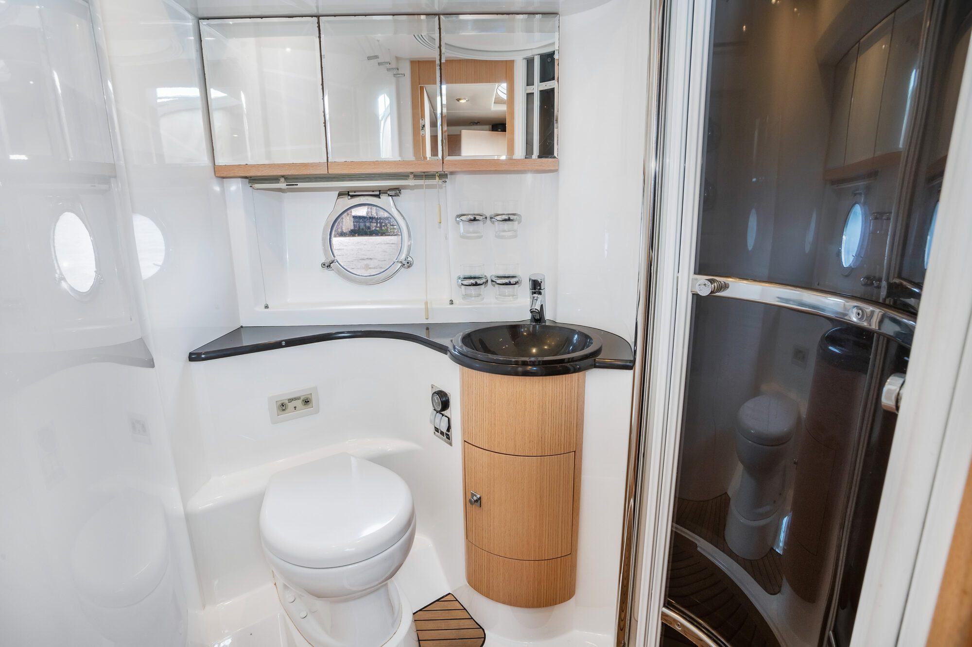 The Birchgrove features a modern yacht bathroom with a round porthole, white walls, a black countertop and sink, a toilet, mirrored cabinets, and a curved glass shower enclosure.