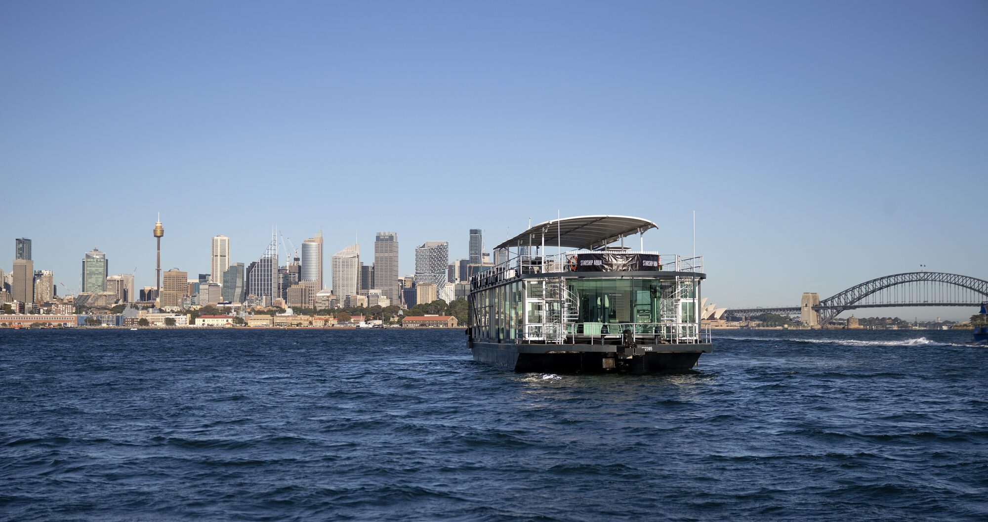 A modern ferry sails on a blue harbor with a city skyline in the background, featuring tall buildings and the Sydney Harbour Bridge under a clear sky.