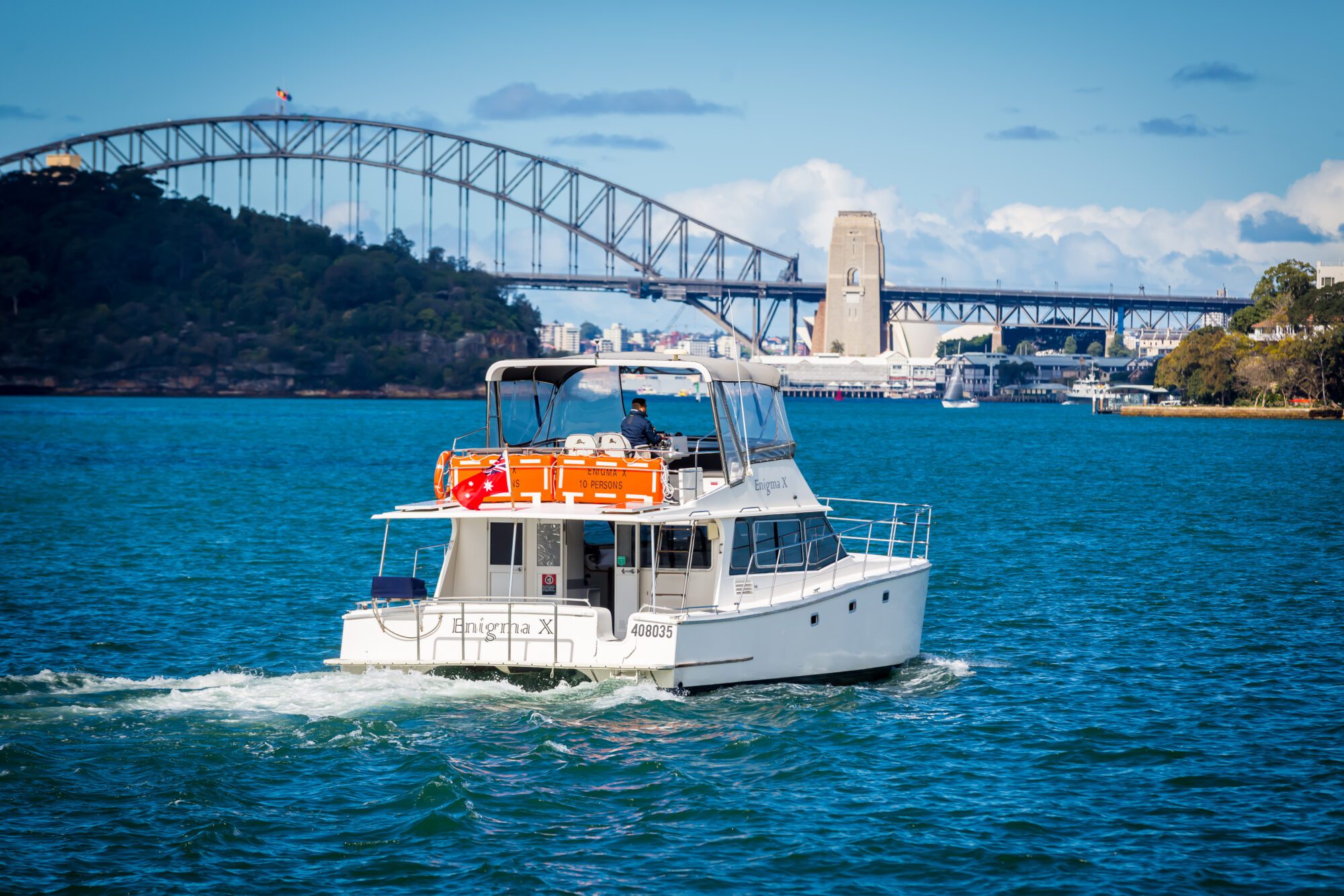A white motorboat cruises on blue water with Sydney Harbour Bridge in the background under a clear sky. The boat has orange life jackets on its roof and people visible on board.