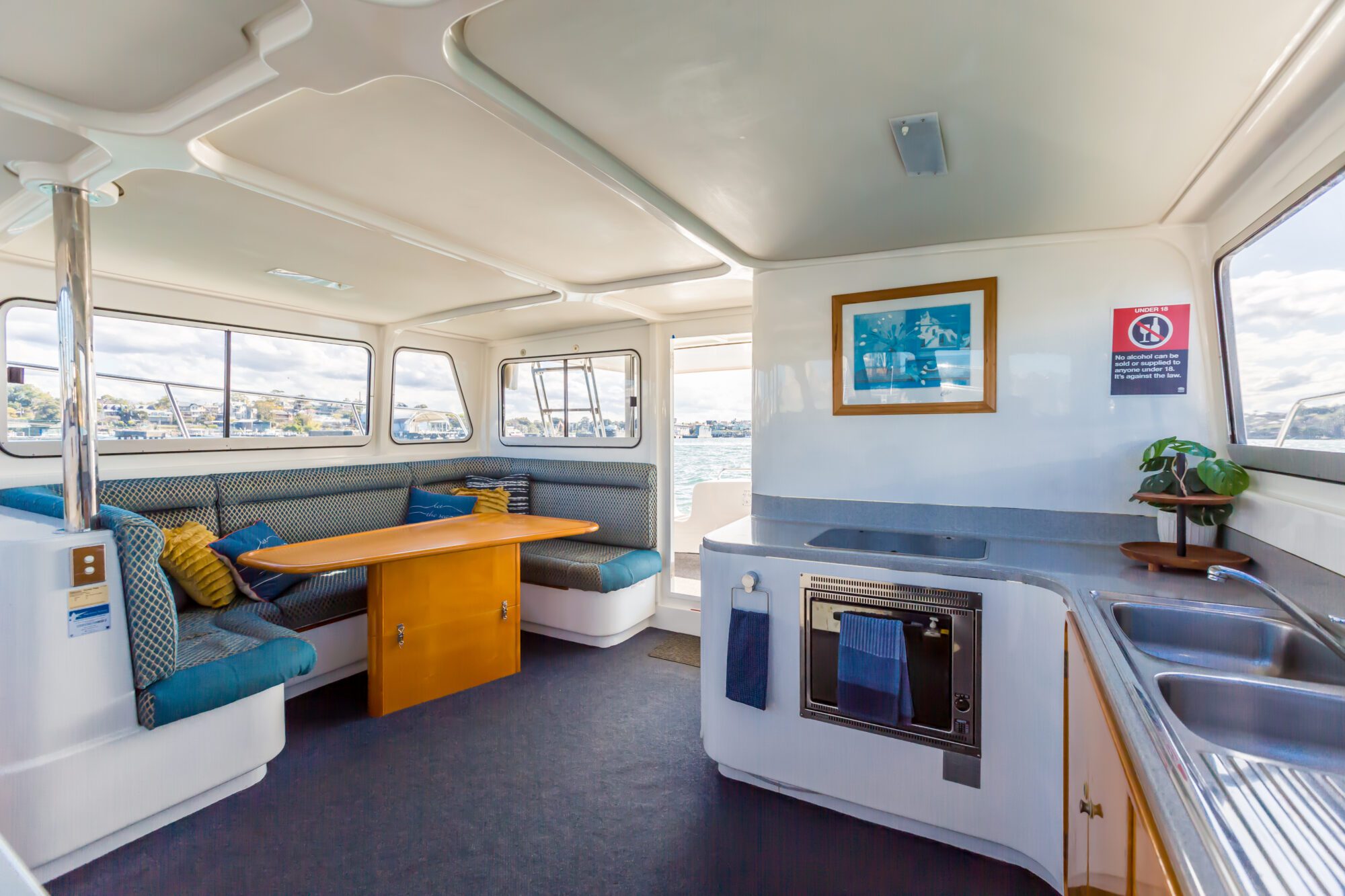 Interior of a boat featuring a dining area with cushioned benches and a wooden table, a kitchenette with a sink, stove, and oven, large windows, and nautical decor. Sunlight streams through the windows.