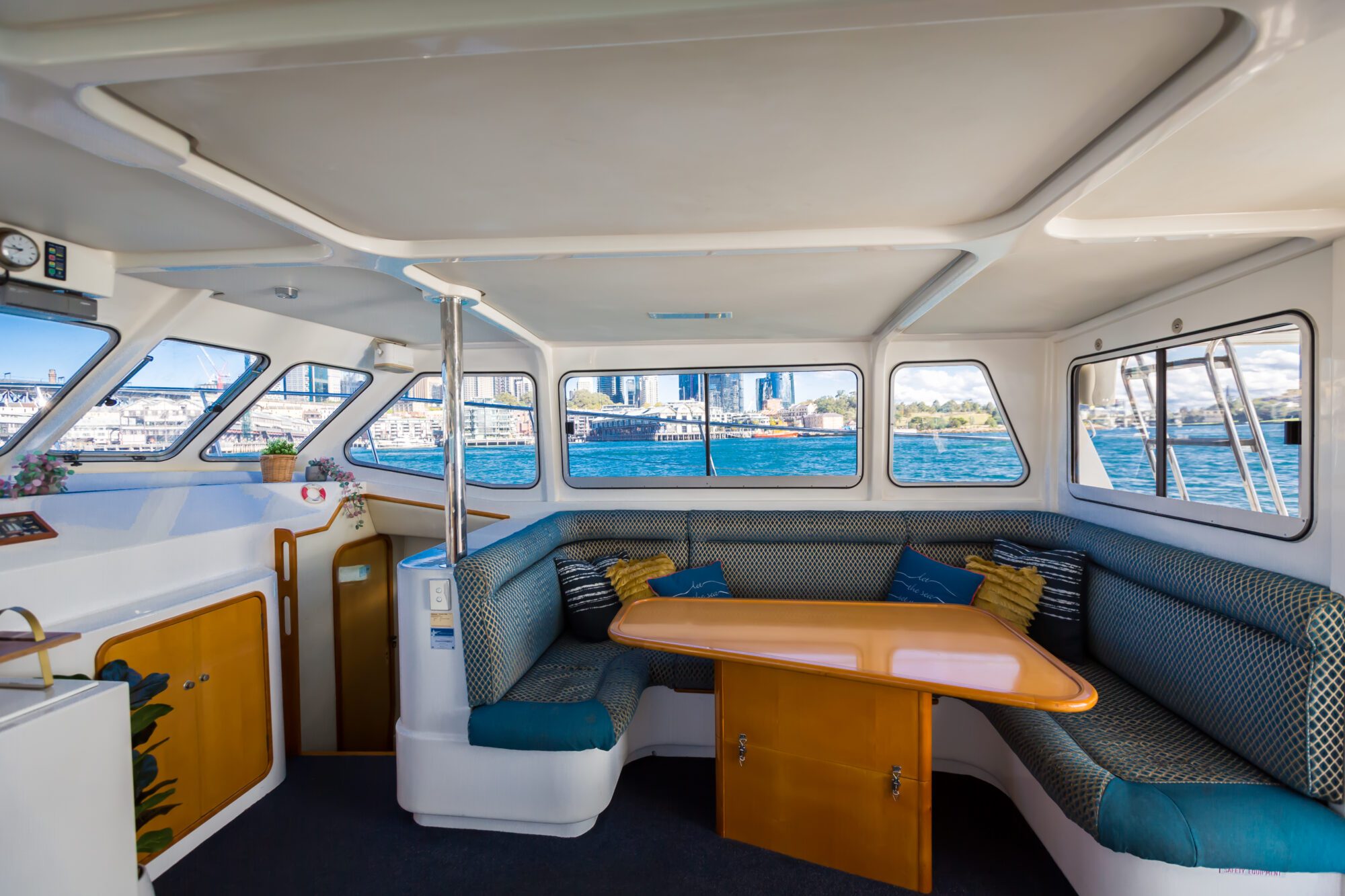 Interior of a boat with a wooden table, cushioned seating, and windows overlooking a city skyline and waterfront, creating a bright and inviting atmosphere for passengers.