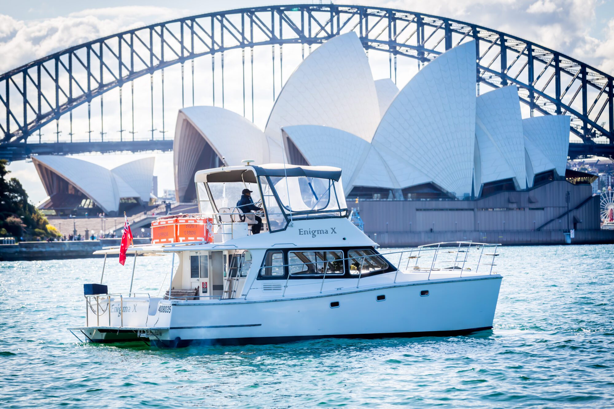 A white yacht named "Enigma X" sails on Sydney Harbour, with the Sydney Opera House and Harbour Bridge visible in the background under a partly cloudy sky.