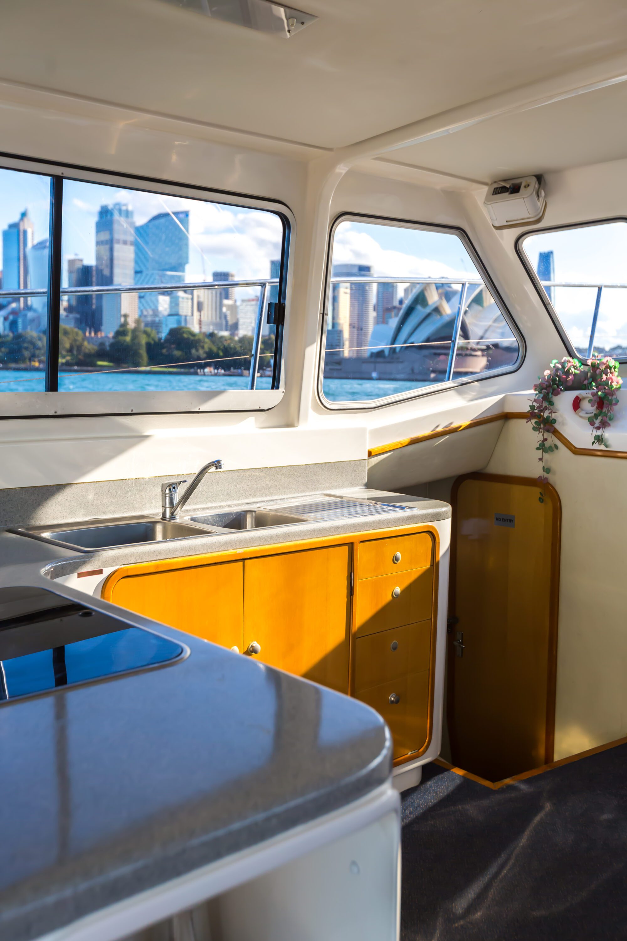 The interior of a boat kitchen features a sink, countertop, wooden cabinets, and a vase of flowers. Large windows provide a view of a city skyline and the Sydney Opera House across the water.