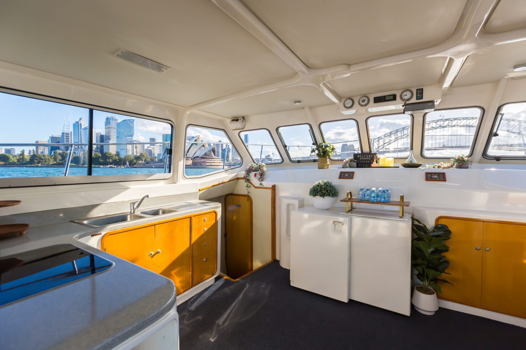 The interior of a boat’s kitchen with yellow and white cabinets, a sink, refrigerator, plants, and bottled water, with large windows showing a city skyline and a bridge over the water outside.
