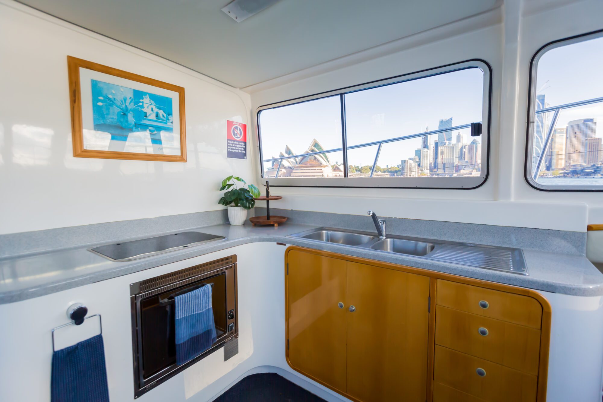 A boat kitchen with a sink, oven, wooden cabinets, a plant, and a framed picture on the wall. Large windows reveal a city skyline and the Sydney Opera House outside.