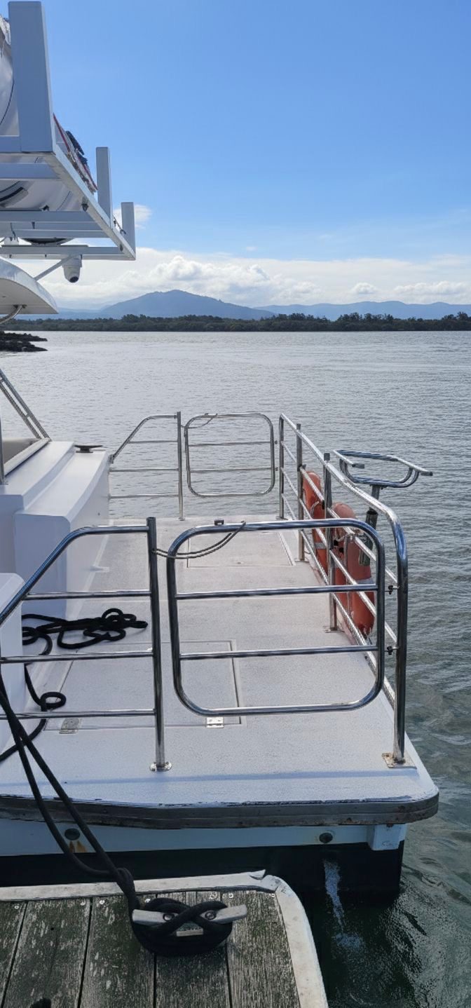 A view from a docked boat showing its railing and an open gate, with calm water, distant hills, and a partly cloudy sky in the background.