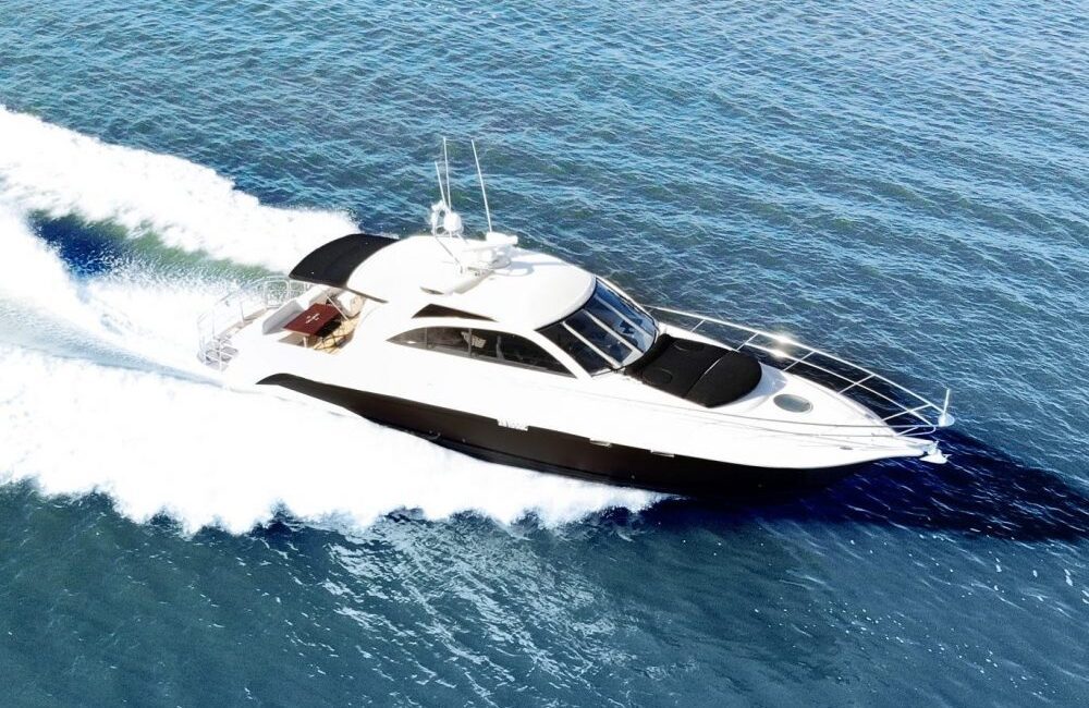 The Coco, a sleek white and black motor yacht, cuts swiftly through clear blue water on a sunny day, leaving a trail of white wake behind.