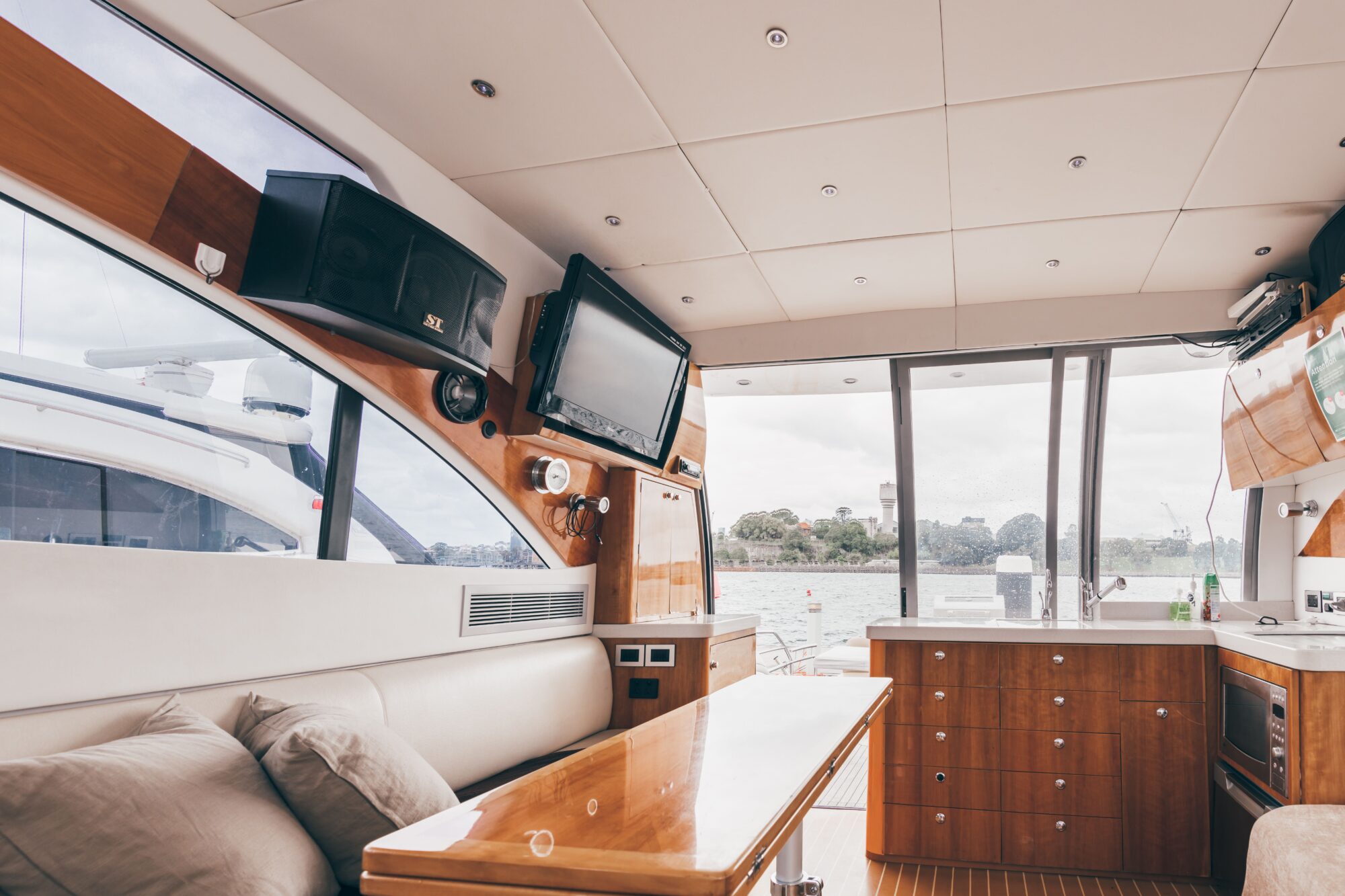 The Coco features a modern boat interior with wood cabinets, a TV, speakers, kitchen area, and large windows offering views of water and trees. Cushioned seating with pillows and a wooden table complete the stylish space.