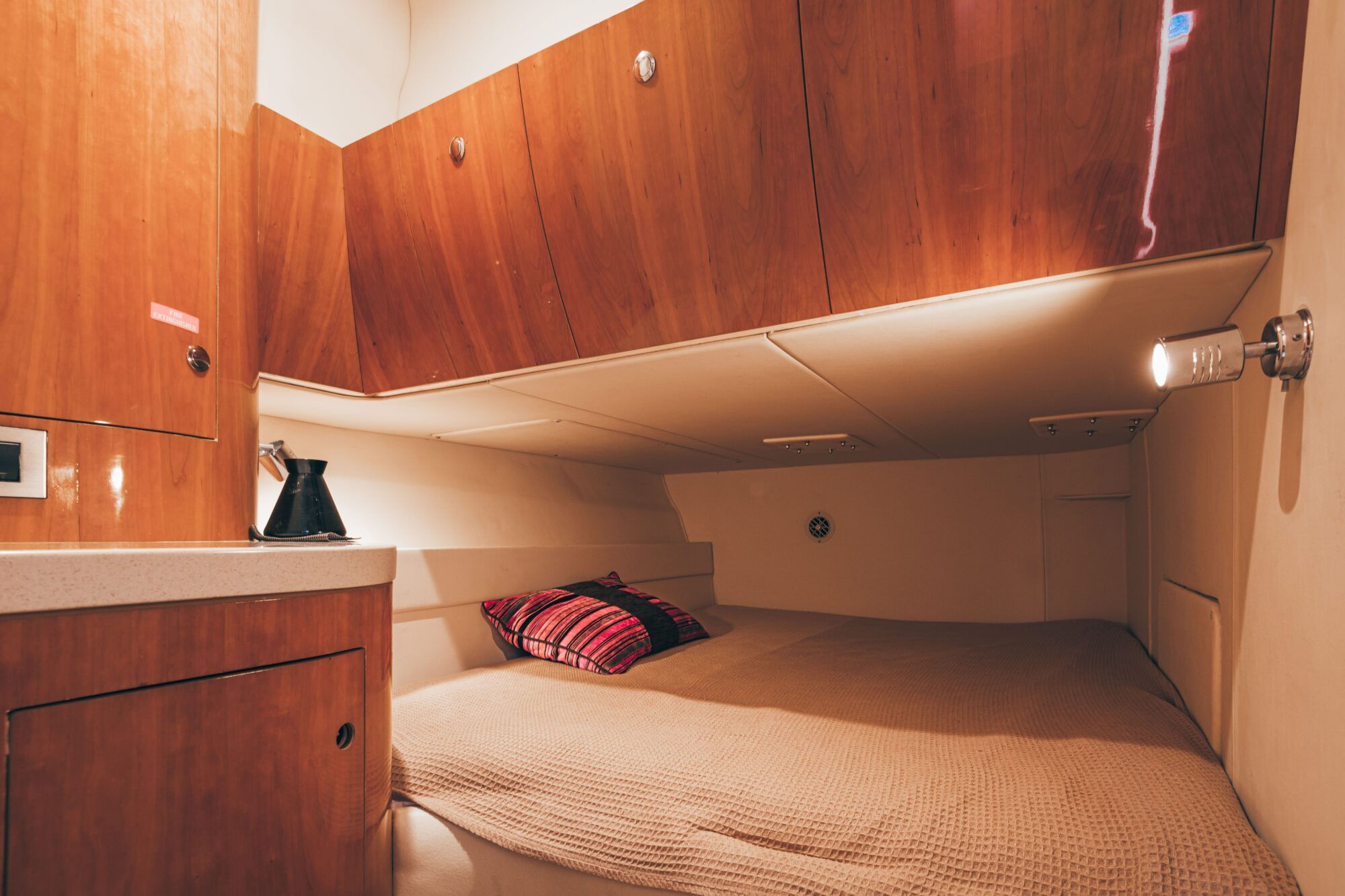 The Coco boat cabin features wooden cabinets, a beige bedspread, two decorative pillows, a compact countertop, and a reading light above the bed, creating a warm and organized atmosphere.
