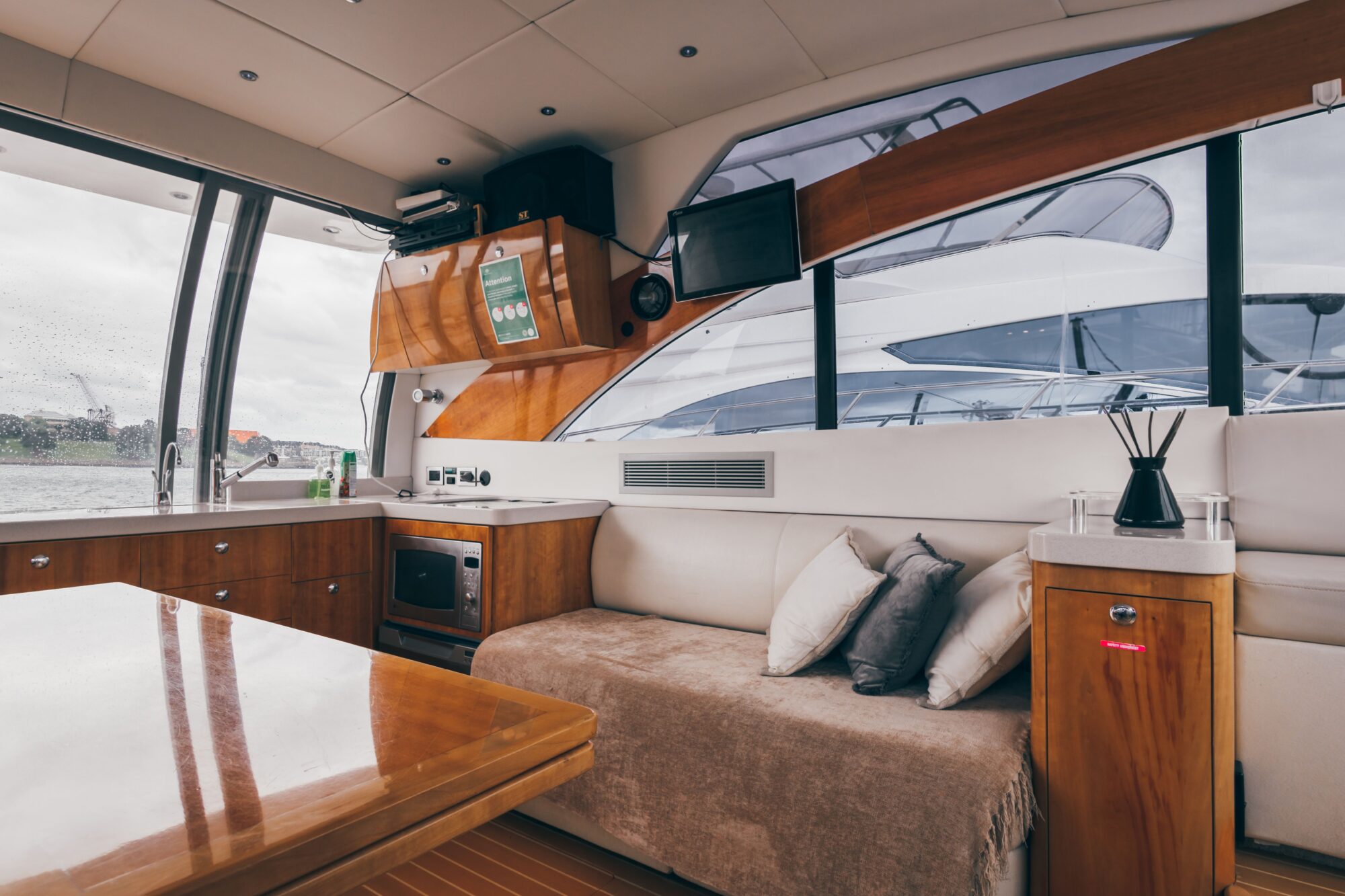The Coco features a modern interior with a wooden table, beige sofa with pillows, compact kitchen, TV, cabinets, and large windows offering views of the water and nearby yachts.