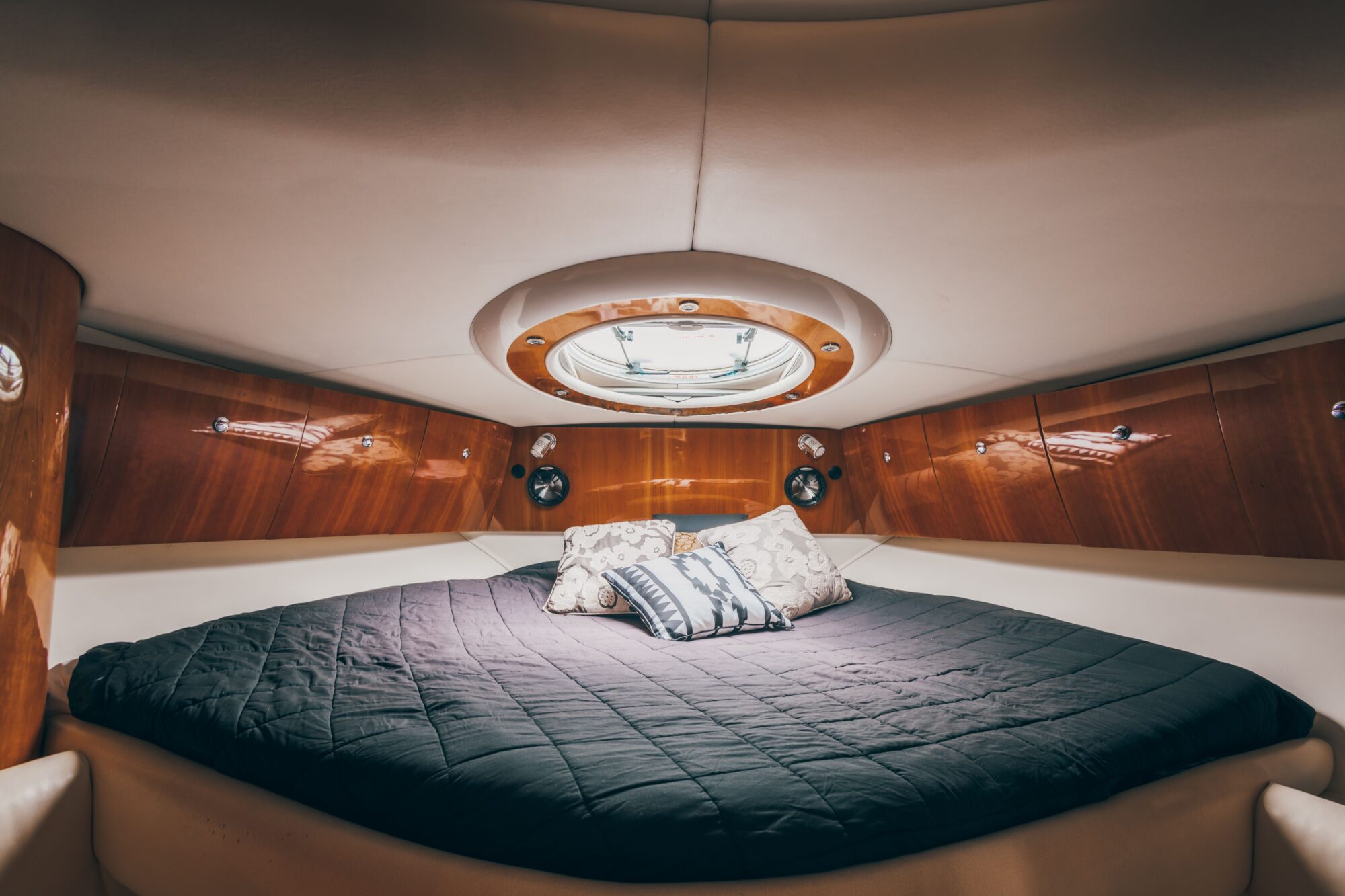 The Coco bed, dressed in dark bedding and decorative pillows, is set in a boat cabin with polished wood paneling and a round overhead window that fills the cozy space with natural light.
