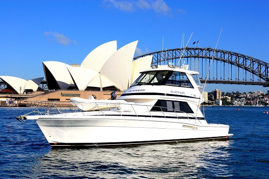 A Platinum yacht glides on the water with the Sydney Opera House and Sydney Harbour Bridge in the background beneath a clear blue sky.