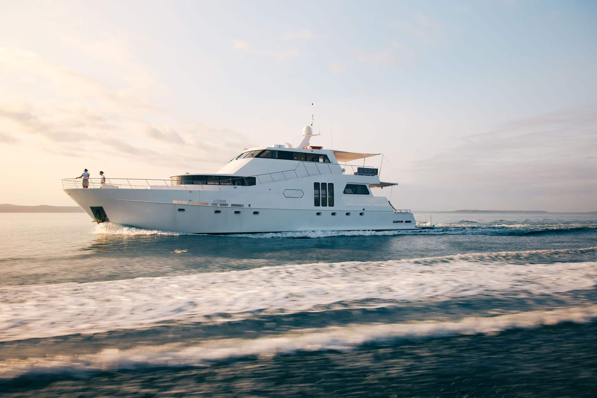 The YOT Viva glides across the water at sunset, leaving a gentle wake. Two people stand at the bow taking in the view, with a partly cloudy sky and a clear horizon stretching out ahead.