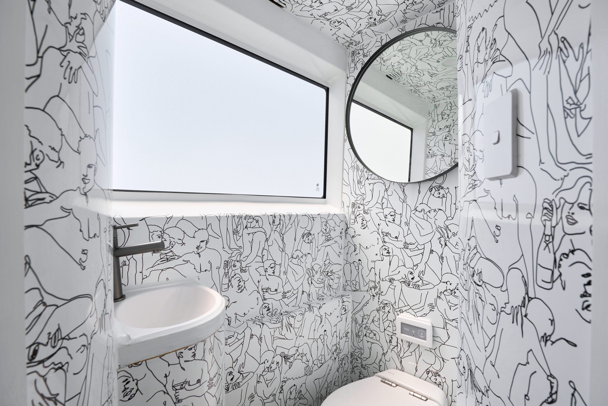 Small modern bathroom with white walls covered in black abstract line art drawings, a round mirror, a small wall-mounted sink, a window, and a toilet.