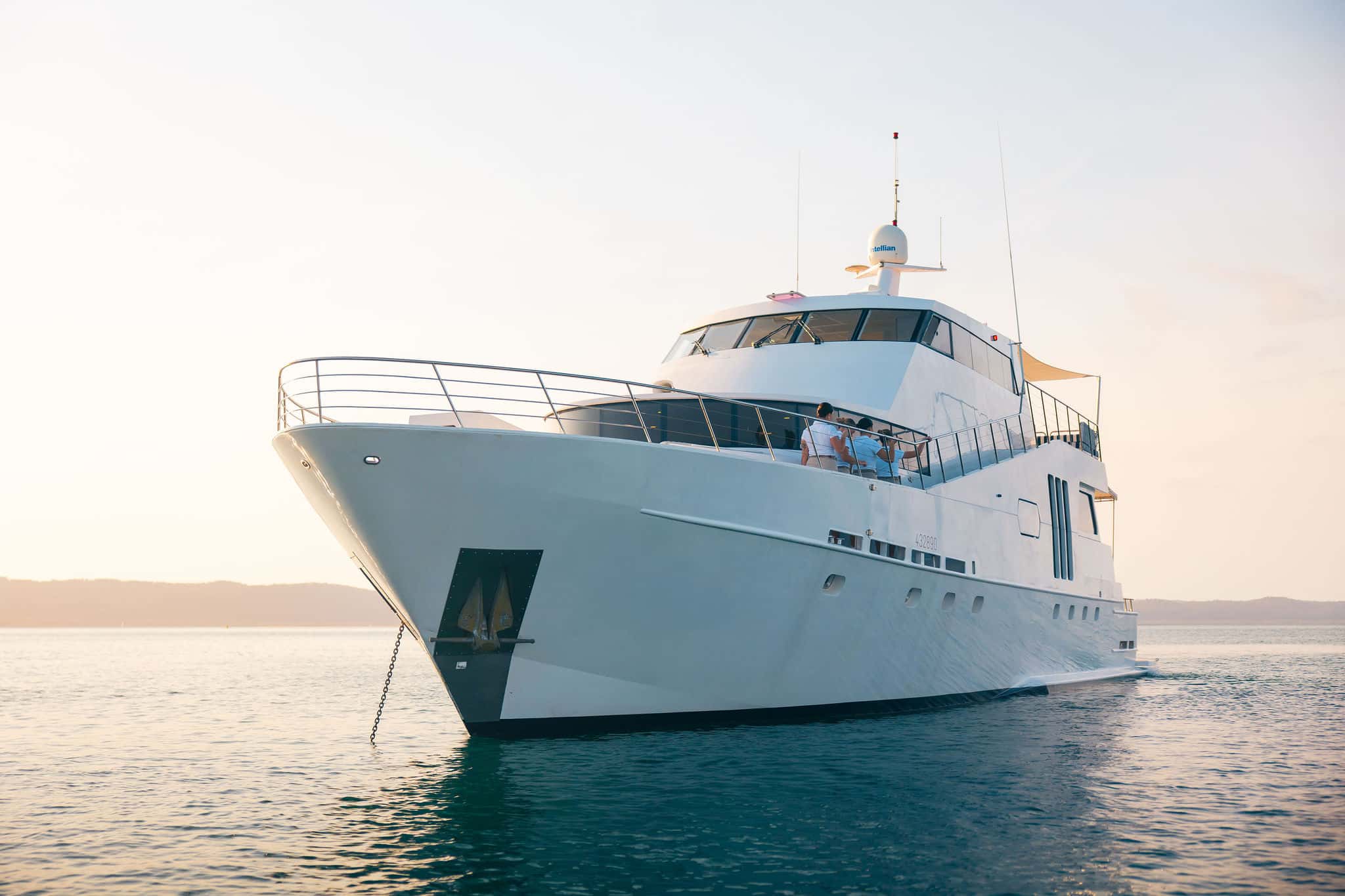 The YOT Viva, a large white luxury yacht, is anchored on calm water at sunset. Two people stand on its deck, with hills visible in the background beneath a clear sky.