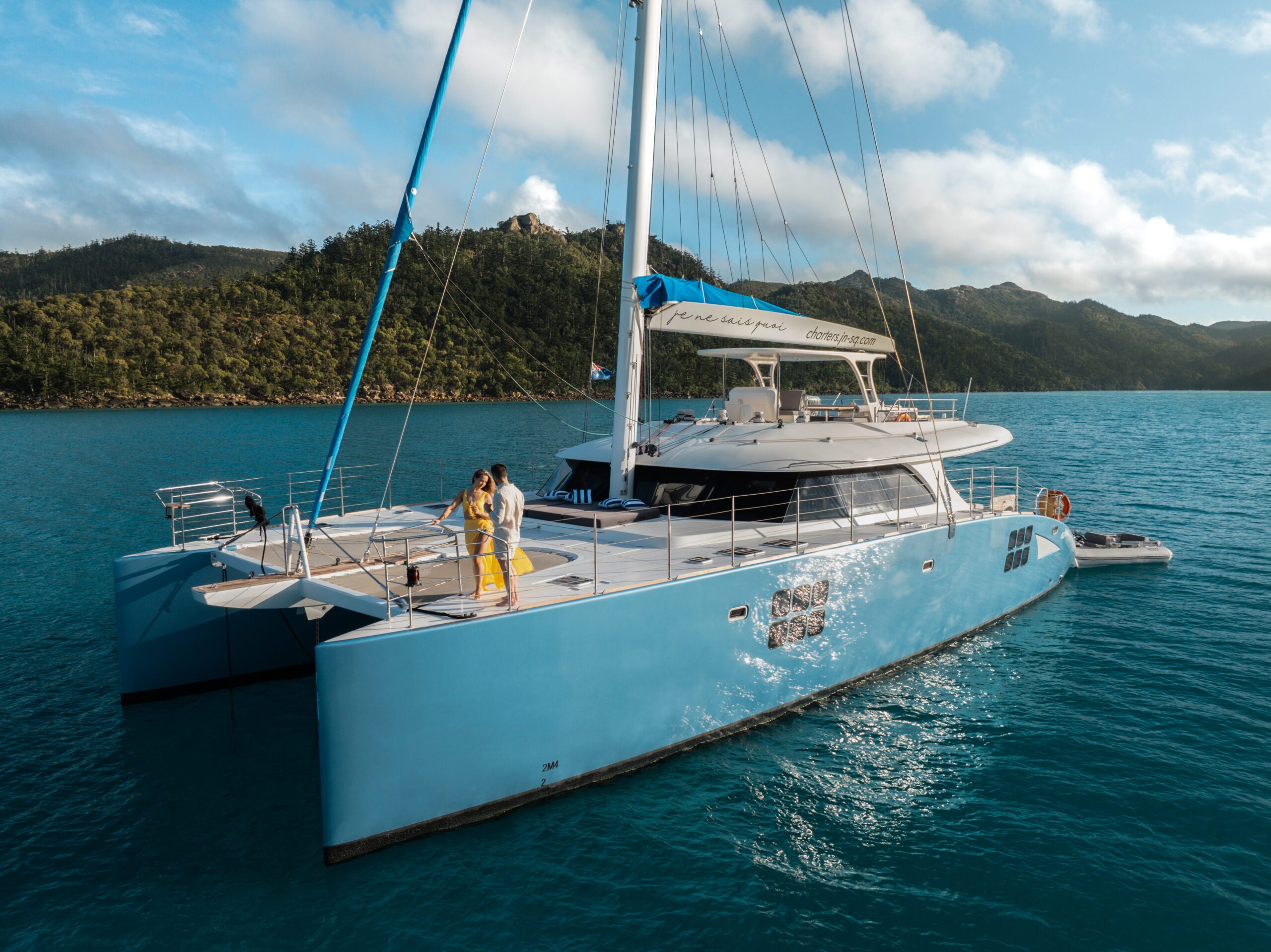 A couple stands on the deck of a large blue catamaran yacht floating on turquoise water, with lush green hills and a partly cloudy sky in the background.