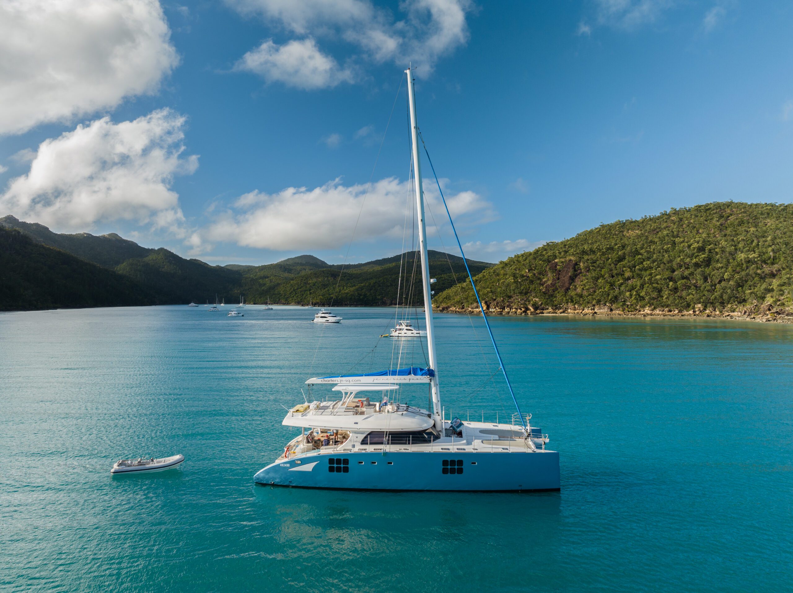 A blue and white catamaran yacht floats on calm turquoise water, surrounded by green hills under a partly cloudy blue sky. Other boats are anchored nearby in the scenic bay.