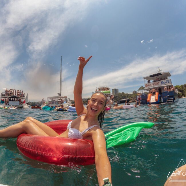 A smiling woman in a white swimsuit floats on a red inflatable in the water, waving at the camera. Boats and people celebrating are visible in the background under a blue sky.
