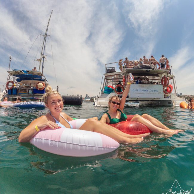 Two women smile and relax on inflatable pool floats in the water near anchored boats, with a crowd enjoying themselves on a large yacht in the background under a bright, partly cloudy sky. “Yacht Social Club” logo in corner.