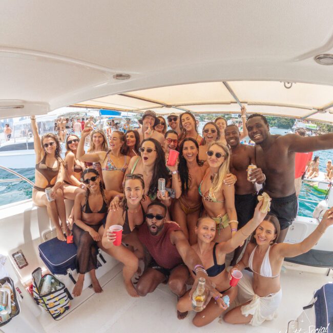 A large group of young adults in swimwear gather on a boat, smiling and holding drinks, enjoying a sunny day on the water with other boats visible in the background.