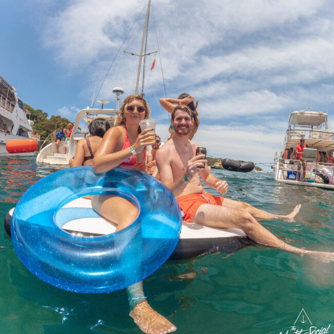 Two people sit on the edge of a boat, smiling and holding drinks, with a blue inflatable ring in front. Other boats and people are visible on the water under a sunny sky. The Yacht Social Club logo is in the corner.
