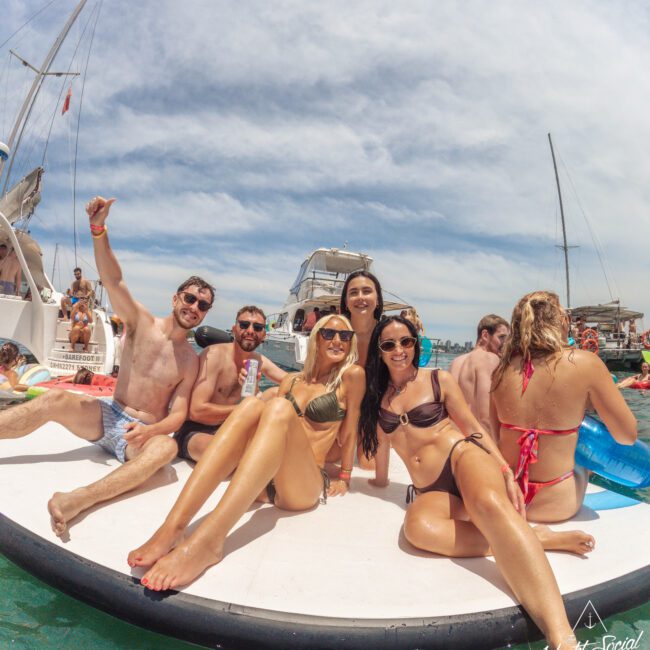 A group of young adults in swimsuits relax and smile on a floating platform in the water, surrounded by boats under a sunny sky, enjoying a lively, social atmosphere.