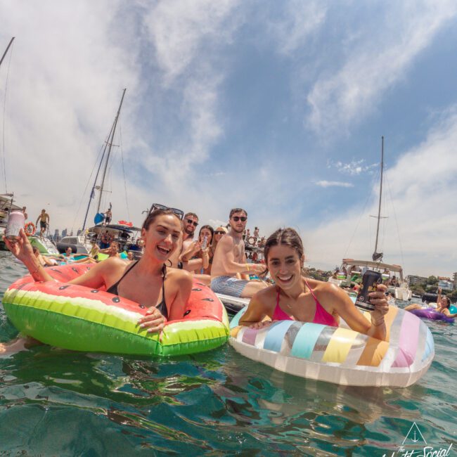 A group of young people relax on colorful pool floats in the sea, smiling and holding drinks, with sailboats and a partly cloudy sky in the background.