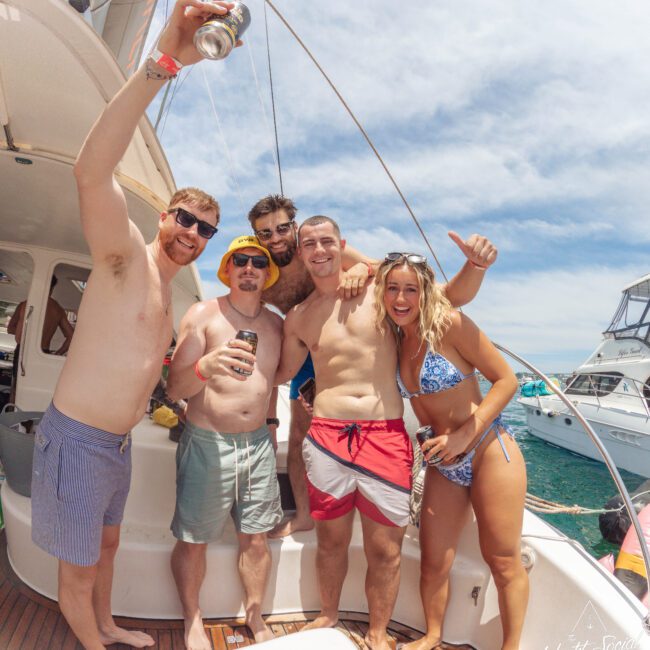 Five friends in swimwear smile and pose together on a boat in sunny weather, holding drinks. Other boats and blue water are seen in the background, suggesting a fun day on the water.
