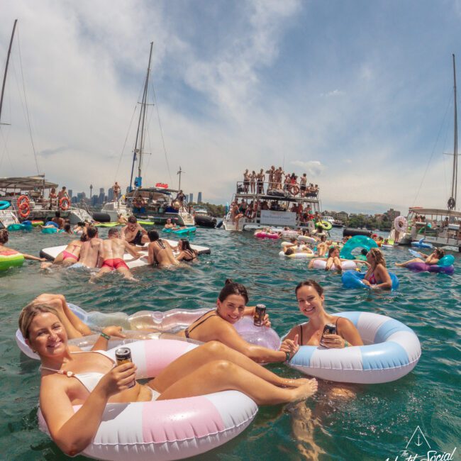 A group of people relax on colorful pool floats in the water, holding drinks, with boats and more partygoers in the background under a partly cloudy sky. The mood is festive and summery.