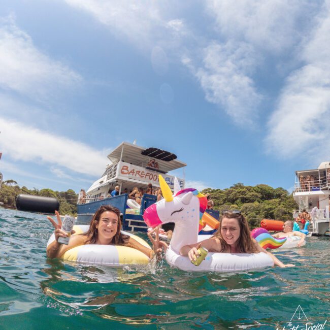 Two women smiling and relaxing on inflatable pool floats, one shaped like a unicorn, in the water near a boat with other people and inflatables, under a blue sky with some clouds.