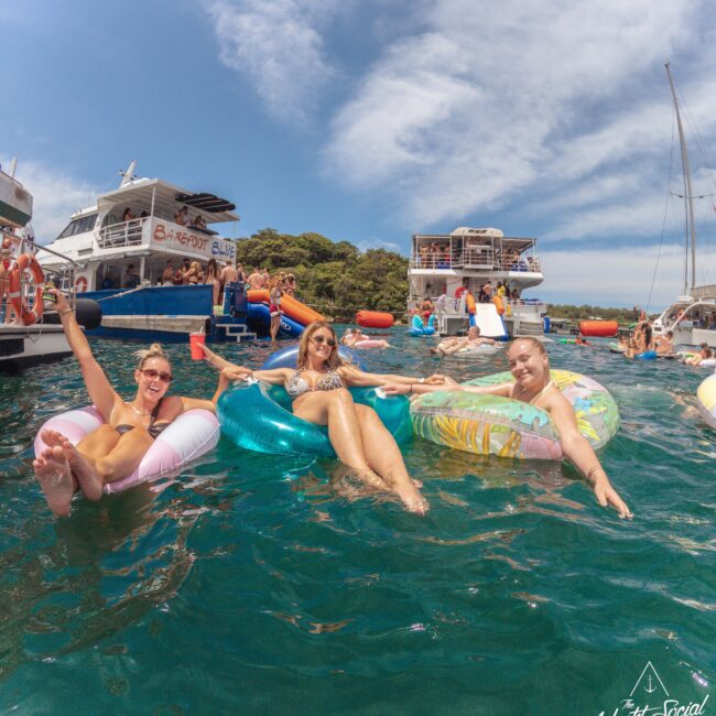Three women relax on inflatable pool floats in clear blue water, surrounded by boats and other people enjoying a sunny day. The scene is lively, with people socializing and celebrating on boats in the background.