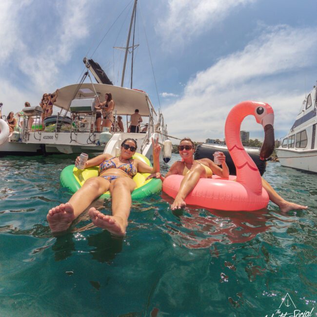 Two women relax on inflatable pool floats, one green and one pink flamingo-shaped, in the water near anchored yachts. They smile, holding drinks, with others socializing on the boats under a sunny sky.