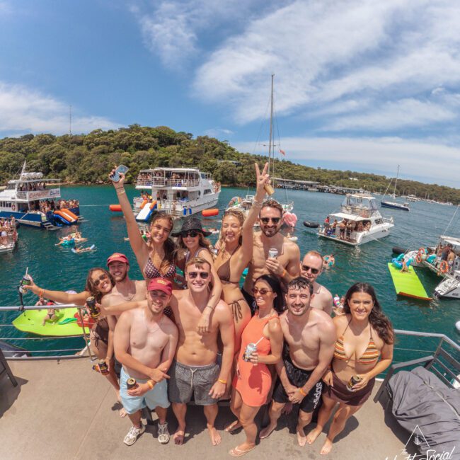 A group of smiling young adults in swimsuits pose together on a boat, holding drinks, with other boats, people swimming, and green trees visible in the background on a sunny day.
