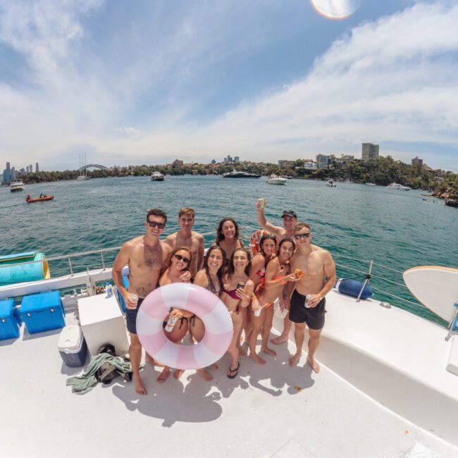 A group of young adults in swimwear pose smiling on a boat, holding drinks and a pink inflatable ring, with a city skyline, boats, and water visible in the background under a sunny sky.