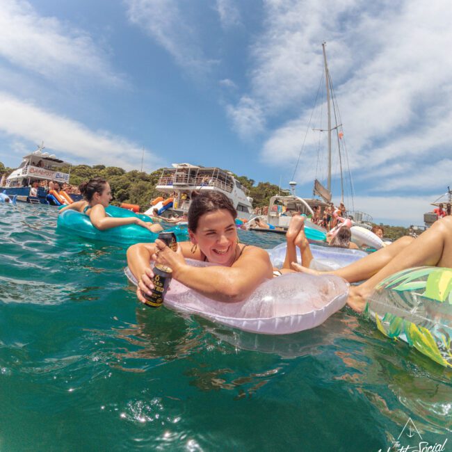 A woman smiles while lounging on a pink inflatable in the water, holding a drink. Other people float nearby on inflatables, with boats and a building in the background under a partly cloudy sky.