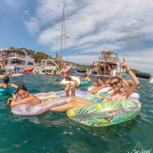 A group of people relax and smile on colorful inflatable floats in the sea, surrounded by boats and yachts under a blue sky. Several people are lounging in the water, enjoying a lively summer social event.