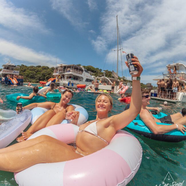 A group of smiling people relax on inflatable floaties in clear blue water, holding drinks, with boats and more partygoers in the background under a sunny sky. The atmosphere is festive and summery.