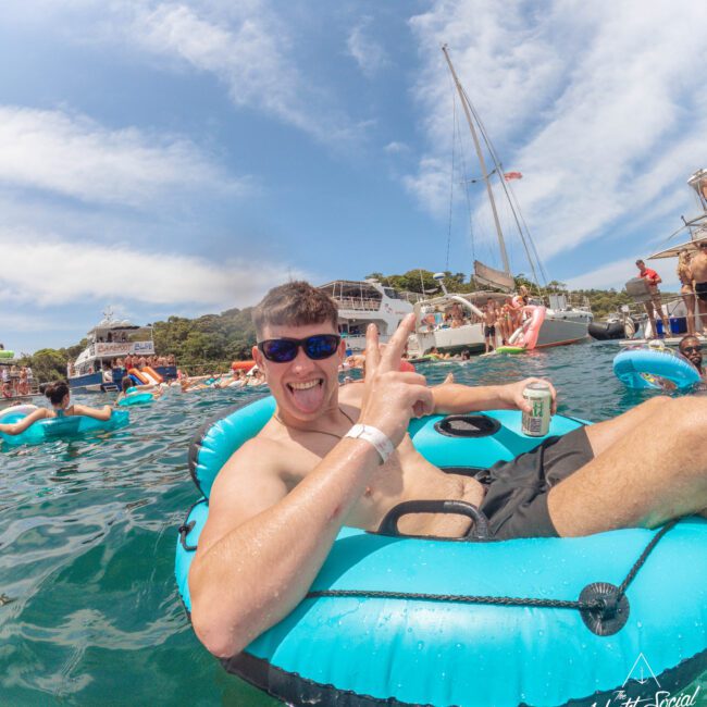 A man in sunglasses smiles and flashes a peace sign while lounging on a blue inflatable in the water, holding a can. Boats and people are in the background under a partly cloudy sky.