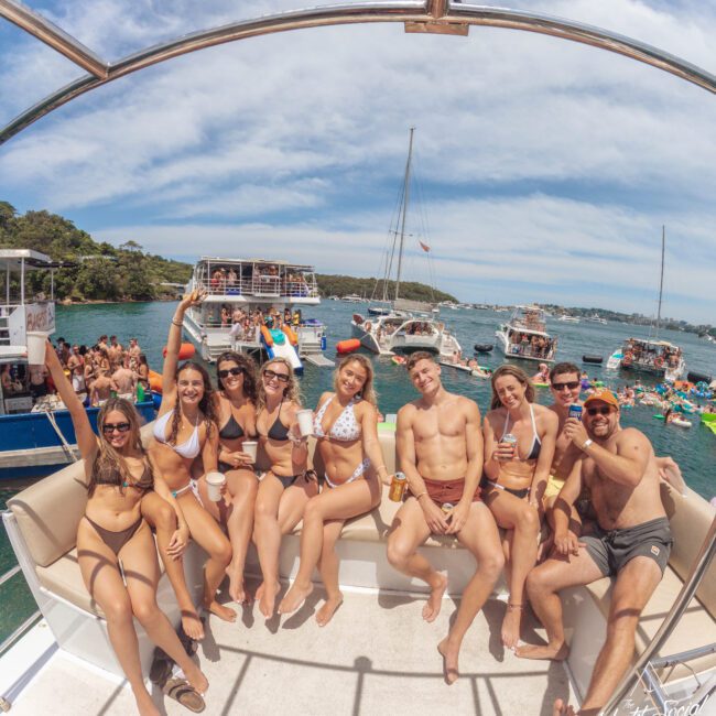A group of young adults in swimsuits relaxes on a boat, smiling and holding drinks, with other boats and people enjoying the sunny day on the water in the background.