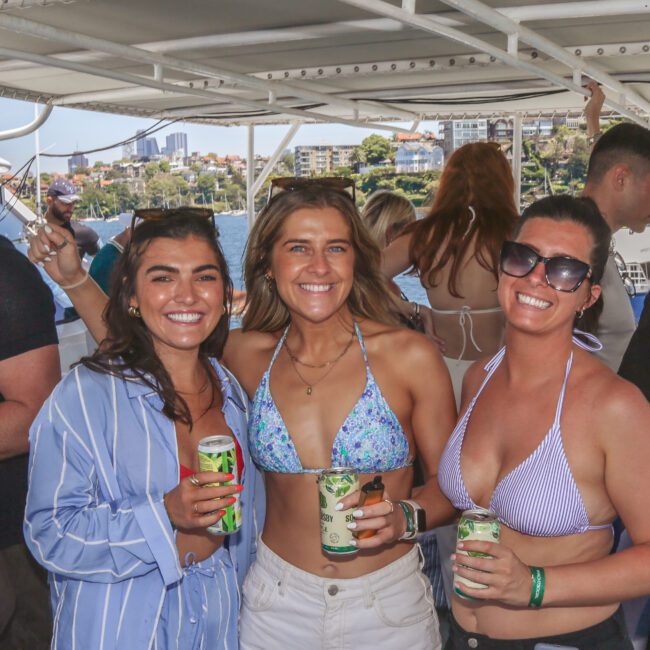 Three women smiling and holding drinks on a boat, dressed in summer outfits with water and city buildings visible in the background. Other people are also seen enjoying the boat party.