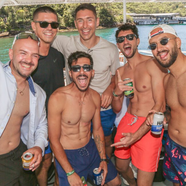 Six men in swimwear and sunglasses smile and pose with drinks on a boat, with clear blue water and lush green trees in the background. The atmosphere is lively and joyful, suggesting a fun summer outing.