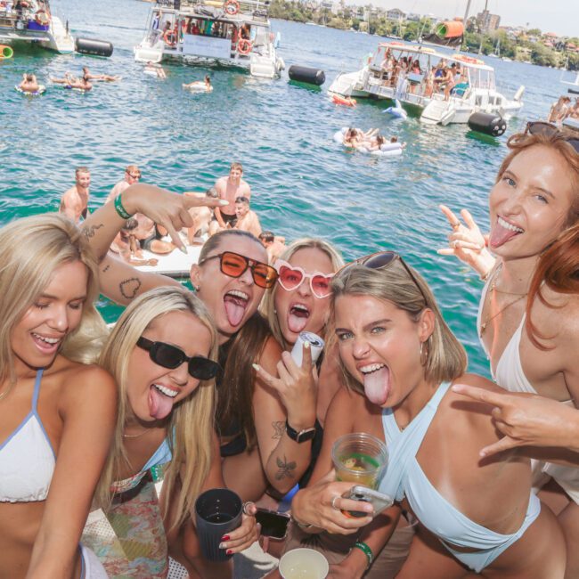Six women in swimsuits smile and pose with their tongues out at a lively lakeside party, surrounded by boats and people swimming in the water on a sunny day.