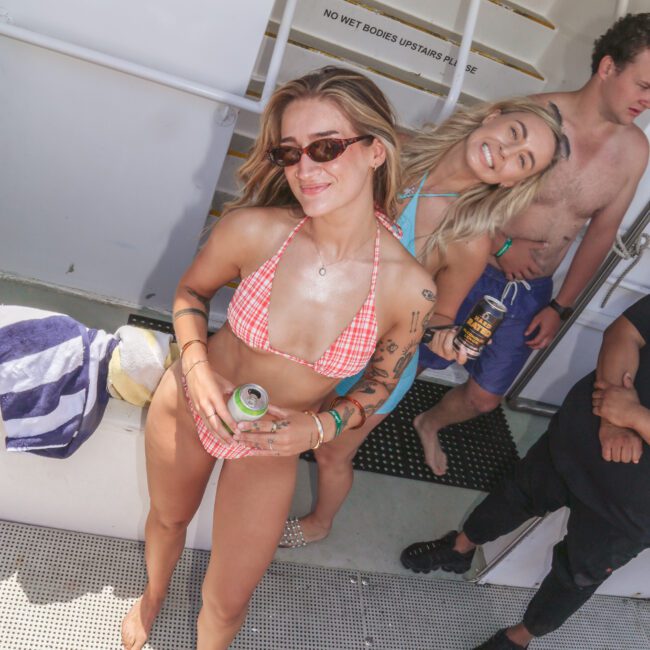 Two women in swimsuits smile at the camera on a boat, one holding a can and wearing sunglasses. Behind them, another woman and a shirtless man stand near a towel and boat railings. Sunlight brightens the scene.