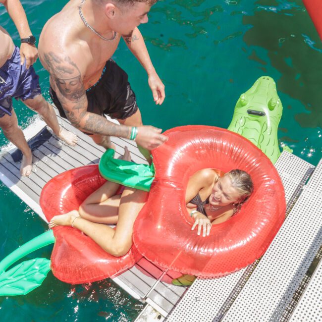 A man helps a woman in a red pool float shaped like a cherry onto a dock by clear blue water. Another person and a green inflatable alligator are nearby. The scene is bright and sunny.