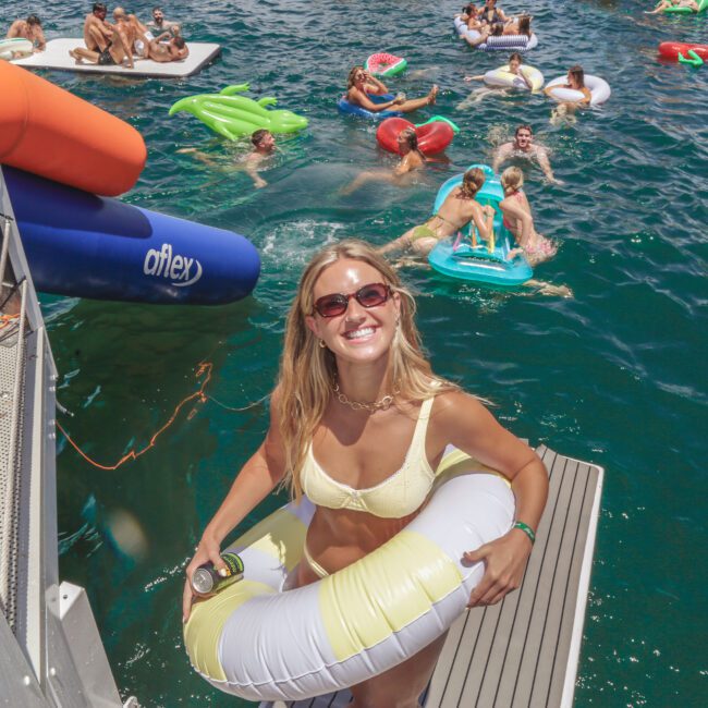 A smiling woman in a yellow bikini and sunglasses holds a pool float and drink on a dock, with people lounging on colorful inflatables in blue water at a lively summer event. "Yacht Social Club" logo is visible in the corner.